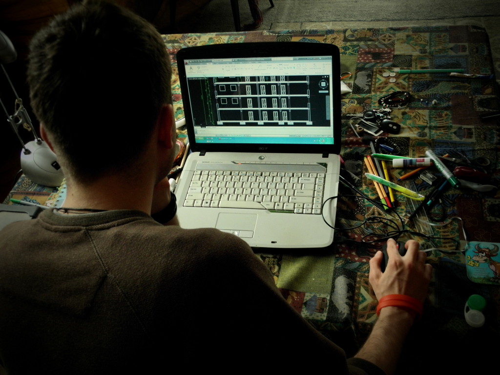 Zdjęcie: Chłopak odwrócony do czytelnika tyłem pracuje na laptopie w programie AutoCad. Na stole leżą kolorowe długopisy
