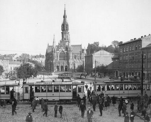 Zdjęcie czarno-białe, stara fotografia przedstawiająca Kraków w dawnych latacg. Na pierwszym planie trawmaj i ludzie, w tle kościoł