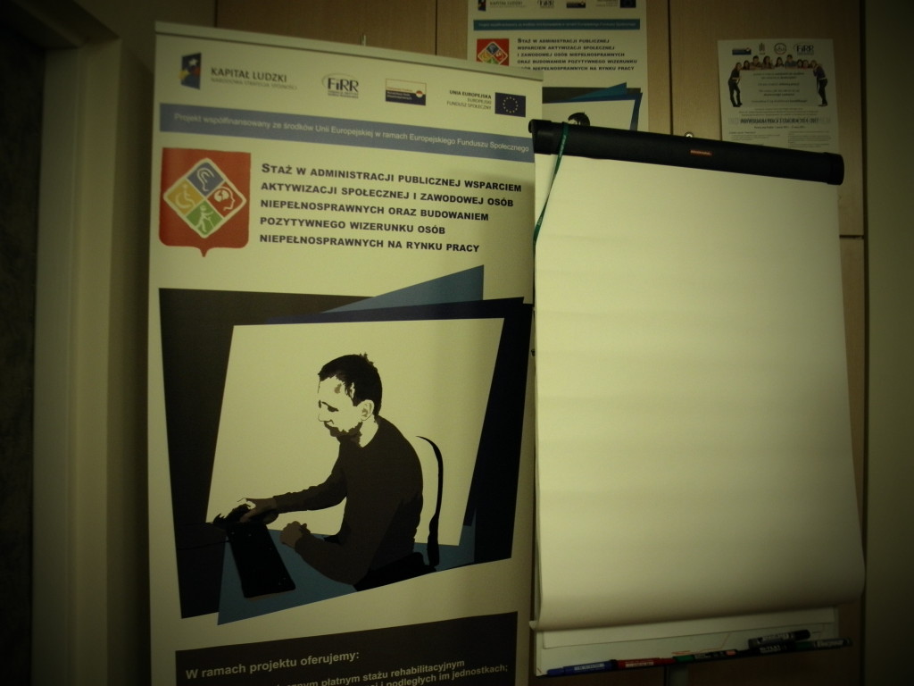 Zdjęcie: Plakat reklamujący projekt stażowy, a obok flipchart z czystymi kartkami w salce szkoleniowej