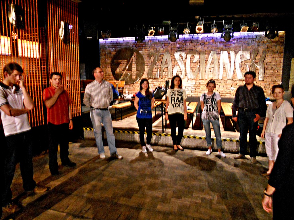 Zdjęcie - na pierwszym planie cztery kobiety oraz czterech mężczyzn stoją w półokręgu, spoglądając ze skupieniem w jednym kierunku. Znajdują się w ciemnym pomieszczeniu rozjaśnionym przez lampy. W tle widać ścianę z napisem 