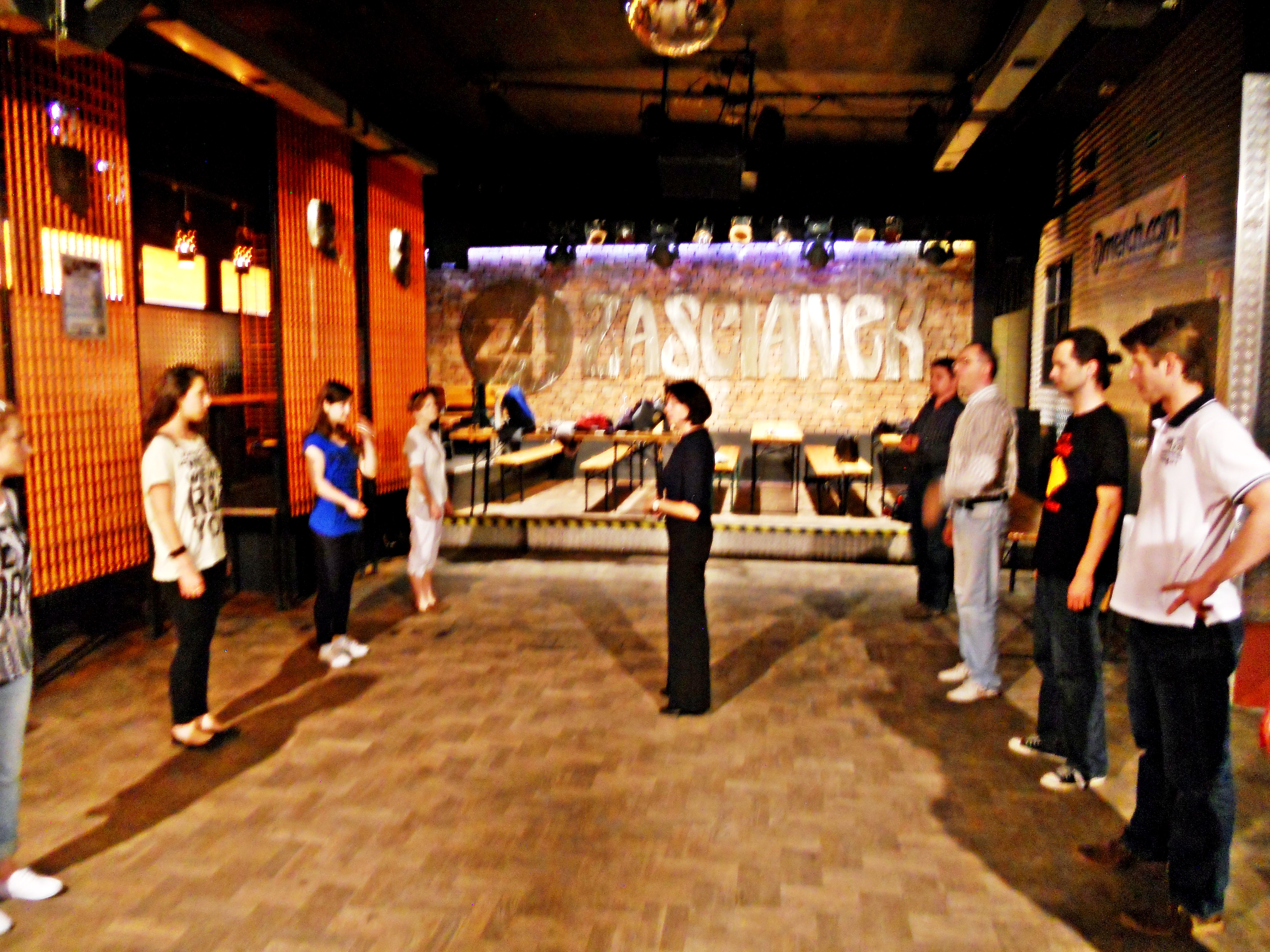 Zdjęcie - na pierwszym planie znajduje się kobieta ubrana na czarno, zwrócona przodem do czwórki innych kobiet i tyłem do czwórki mężczyzn. W tyle na ścianie widnieje napis 